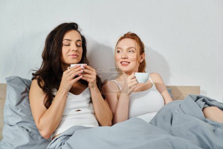 Téléchargez les photos : Deux femmes en tenue élégante s'assoient sur un lit, souriant tout en tenant des tasses à café. - en image libre de droit