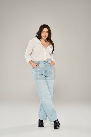 A beautiful plus size woman strikes a pose in a white shirt and blue jeans against a gray backdrop.