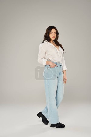 Stylish plus size woman posing confidently in white shirt and blue jeans on a gray backdrop.