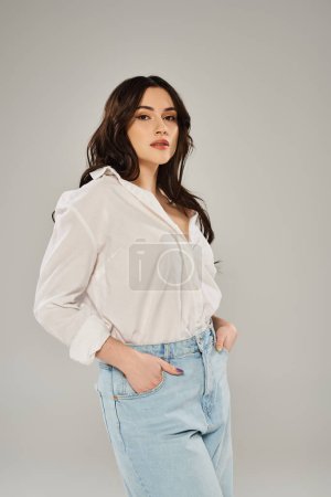 A beautiful plus size woman striking a pose in a white shirt and jeans against a gray backdrop.