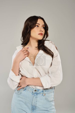 A beautiful plus size woman confidently poses in a white shirt and jeans against a gray backdrop.