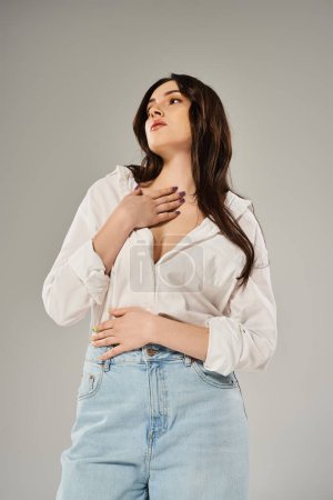 A stunning plus size woman strikes a pose in a trendy white shirt and blue jeans against a neutral gray backdrop.