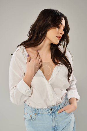 Téléchargez les photos : A beautiful plus-size woman poses confidently in a white shirt and jeans against a gray backdrop. - en image libre de droit