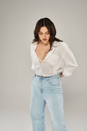 A beautiful plus size woman poses confidently in a stylish white shirt and blue jeans against a gray backdrop.