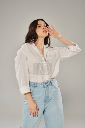 A beautiful plus size woman poses confidently in a white shirt and jeans against a gray backdrop.