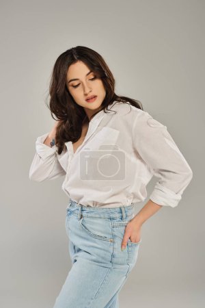 A stunning plus size woman poses in a trendy white shirt and jeans against a gray backdrop.