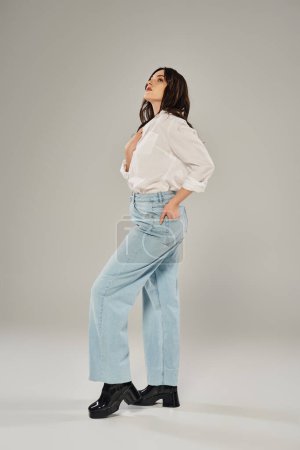 A plus size woman exudes confidence in a white shirt and blue jeans against a gray backdrop.