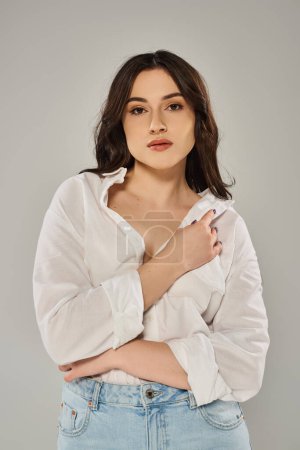 Stylish plus-size woman posing in trendy white shirt and jeans against a gray backdrop.