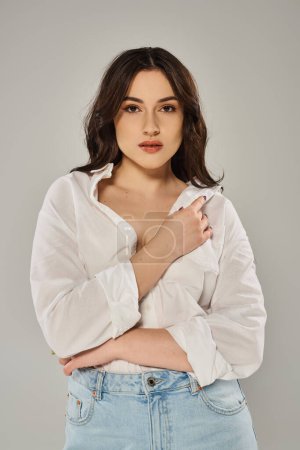 Una impresionante mujer de talla grande posa en una camisa blanca sobre un fondo gris, exudando confianza y estilo.