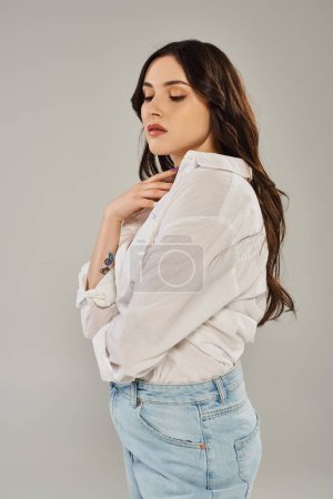 A stunning plus-size woman poses elegantly in a white shirt and jeans against a gray backdrop.