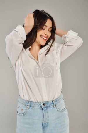 A beautiful plus size woman posing in stylish white shirt and jeans against a gray backdrop.