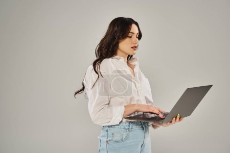 A beautiful plus size woman in stylish attire holds a laptop with confidence on a gray backdrop.