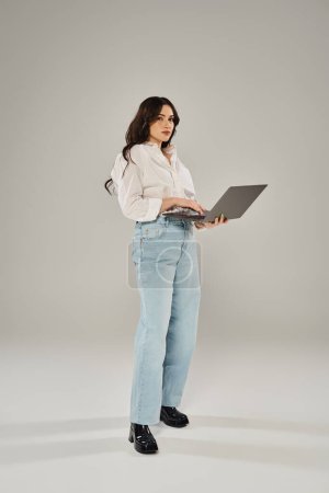 Plus-Size-Frau steht selbstbewusst mit Laptop in schicker Kleidung vor grauem Hintergrund.