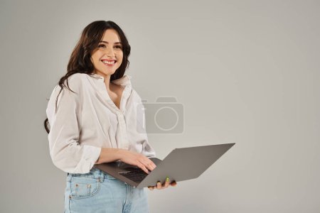 Foto de Una mujer elegante de tamaño grande sonríe mientras sostiene una computadora portátil contra un telón de fondo gris. - Imagen libre de derechos