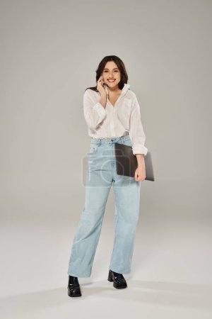 Plus size woman in white shirt and jeans posing confidently with a laptop on a gray backdrop.