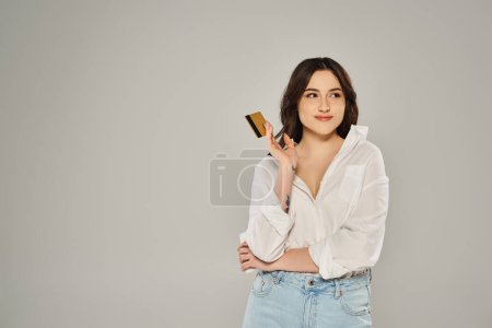 Una elegante mujer de talla grande con una camisa blanca posando mientras sostiene un teléfono celular contra un telón de fondo gris.