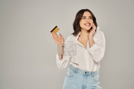 Una mujer elegante de tamaño grande multitarea, sosteniendo una tarjeta de crédito y hablando en un teléfono celular contra un fondo gris.