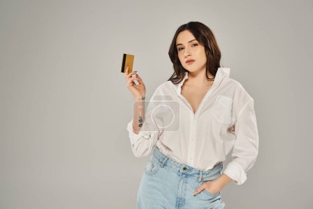 Beautiful plus-size woman in a white shirt confidently holding a gold card against a gray backdrop.