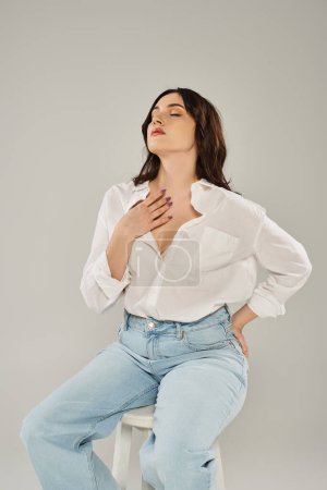 A beautiful plus size woman in stylish attire sitting on a stool, gracefully placing her hands on her chest.