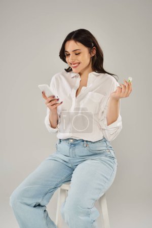 A plus-size woman in stylish attire engrossed in using a cellphone while sitting on a stool against a gray backdrop.