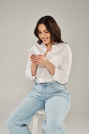 Eine modische Plus-Size-Frau, stilvoll gekleidet, sitzt auf einem Schemel, während sie ein Handy benutzt.