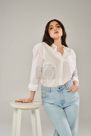 Photo for A beautiful plus-size woman in stylish attire confidently sits atop a stool against a gray backdrop. - Royalty Free Image