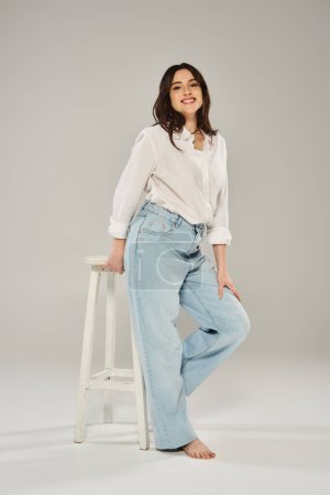 A beautiful plus size woman poses confidently in a white shirt and jeans on a gray backdrop.