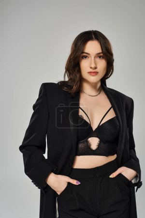 Eine schöne Plus-Size-Frau posiert selbstbewusst in einem eleganten schwarzen Anzug und BH vor grauem Hintergrund.