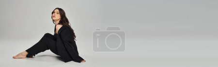 A beautiful plus-size woman sits gracefully on the ground in a stylish black outfit against a gray backdrop.
