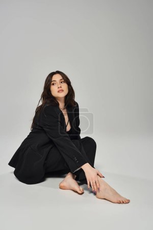 A beautiful plus-size woman in stylish attire sits gracefully with her legs crossed on a gray backdrop.