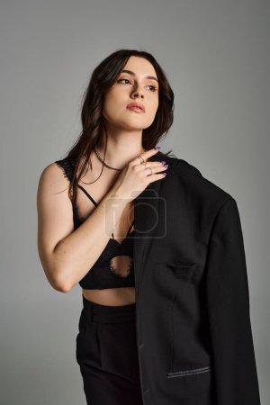 Stylish plus size woman confidently poses in a black suit and bra against a gray backdrop.