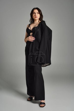 Plus-size woman exuding elegance in a black dress and shawl against a gray backdrop.