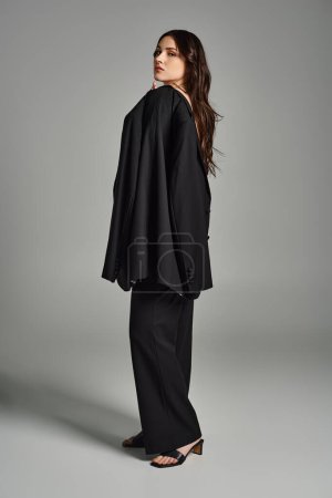 Stylish plus size woman confidently posing in a black suit and heels on a gray backdrop.