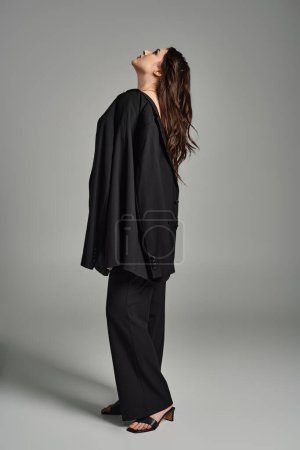 Foto de Una hermosa mujer de talla grande toma una pose en un elegante traje negro y tacones contra un fondo gris. - Imagen libre de derechos