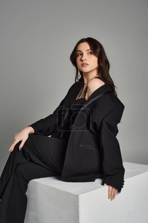 A beautiful plus size woman in stylish attire sitting regally on a white block against a gray backdrop.