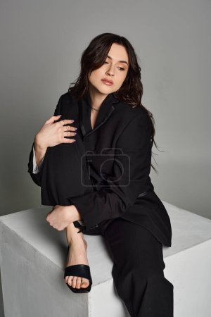 A plus size woman in stylish attire sits gracefully on a white block against a gray backdrop, exuding elegance and confidence.