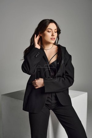 Plus size woman exuding confidence in a sleek black suit striking a pose on a gray backdrop.