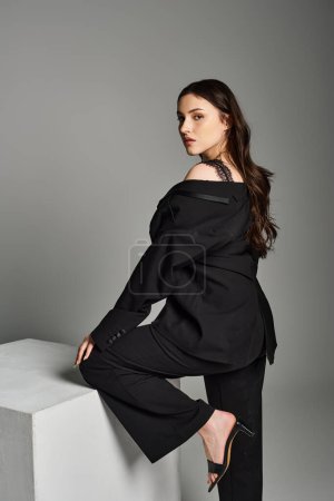 Eine atemberaubende Plus-Size-Frau präsentiert ihren Stil in einem schwarzen Top und einer schwarzen Hose vor grauem Hintergrund.