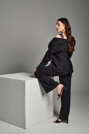Eine schöne Plus-Size-Frau posiert selbstbewusst in schwarzem Top und Hose vor grauem Hintergrund.