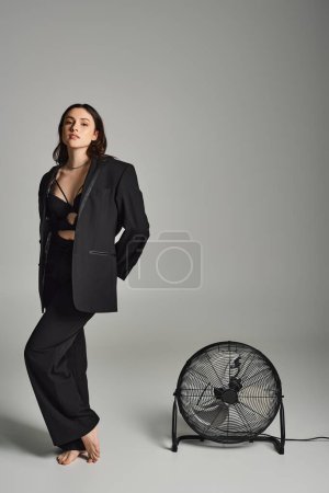 Téléchargez les photos : A beautiful plus size woman in a black suit standing gracefully next to a fan on a gray backdrop. - en image libre de droit
