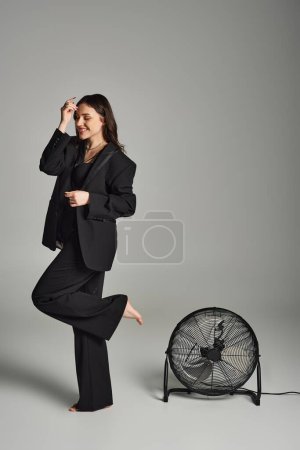 Una hermosa mujer de talla grande con un atuendo elegante, de pie con gracia delante de un ventilador, su traje revoloteando en la brisa.