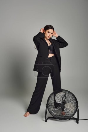 A plus-size woman exudes confidence in a stylish suit, standing gracefully next to a rotating fan against a gray backdrop.