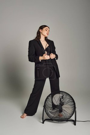 Eine stylische Plus-Size-Frau strahlt in einem maßgeschneiderten Anzug Zuversicht aus und steht anmutig neben einem sich drehenden Fächer vor grauem Hintergrund..