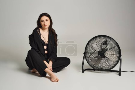 Une belle femme de grande taille en tenue élégante s'assoit gracieusement à côté d'un ventilateur tournant sur un fond gris.