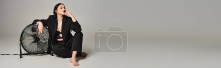Eine stylische Plus-Size-Frau sitzt neben einem Ventilator und genießt einen erfrischenden Moment vor grauem Hintergrund.