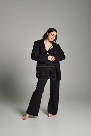 Plus size woman exudes confidence in a black suit against a gray backdrop while posing for the camera.