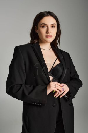 Eine schöne Plus-Size-Frau posiert selbstbewusst in einem eleganten schwarzen Anzug vor grauem Hintergrund und strahlt Kraft und Eleganz aus.