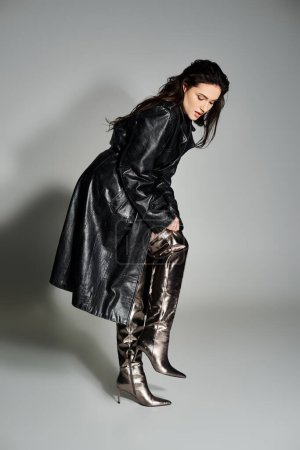 A beautiful plus size woman poses in a stylish black coat and boots against a gray backdrop.