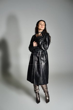 Eine atemberaubende Plus-Size-Frau posiert in schickem schwarzen Mantel und Stiefeln vor grauem Hintergrund.