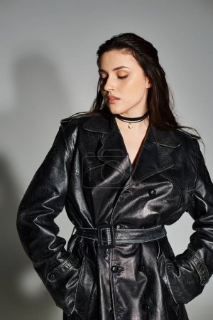 A beautiful plus-size woman strikes a confident pose in a stylish black leather trench coat against a neutral gray backdrop.